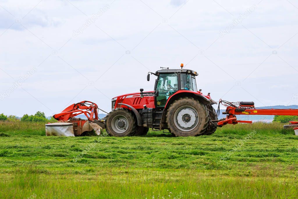 A big red tractor mows the grass for silage on the field
