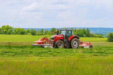 İki çim biçme makinesi olan büyük kırmızı bir traktör bir silonun üzerindeki yeşil çimleri biçiyor..