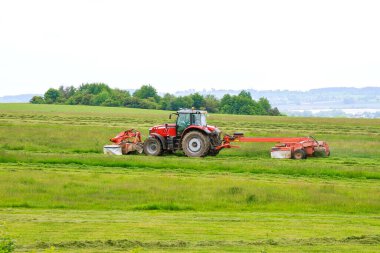 Kırmızı bir traktör iki çim biçme makinesiyle çimleri biçer. Kış için hayvanlar için saman topluyorum..