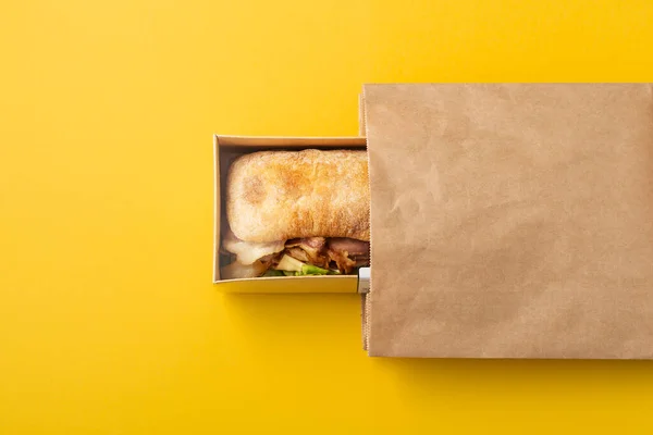 Sandwich in takeout or delivery food container and paper bag on yellow background, top view