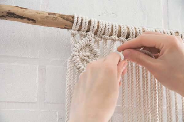 женщина вяжет плетение макраме на деревянной палочке рядом с белой кирпичной стеной