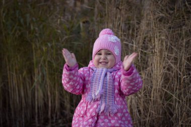 Pembe ceketli ve şapkalı küçük bir kız mutlu bir şekilde gülümseyip ellerini çırpıyor. Güneşli bir sonbahar gününde kuru kamışların arka planında pembe elbiseli güzel bir kız..