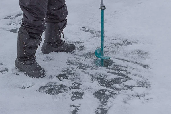 A man drills a hole with an ice screw close-up. Winter fishing. A fisherman drills a hole in the ice for ice fishing. Male legs in boots and ice screw on ice background