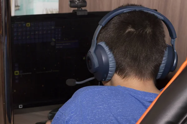 A parte de trás da cabeça de um adolescente com auscultadores azuis atrás do monitor. Um rapaz com auscultadores estuda em casa. Um jogador adolescente joga no computador com fones de ouvido. Visão traseira — Fotografia de Stock