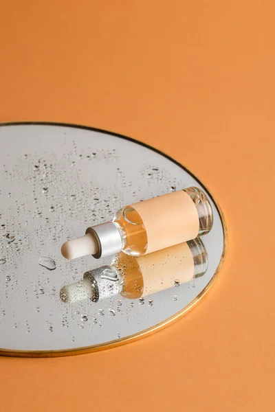 Açık turuncu arka planda damlaları olan bir aynada deri bakımı için pipeti olan şeffaf bir serum şişesi. Sağlık ve güzellik kavramı.