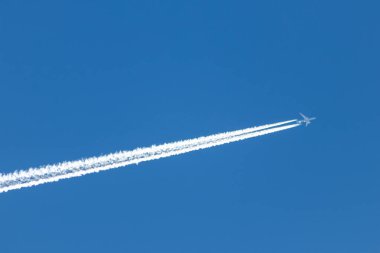 Mavi gökyüzünde uzun, beyaz, dumanlı egzoz dumanı ve jet yıkamasıyla uçan uçak, pilotlarla uluslararası ulaşım ve küreselleşme tatillerini gösteriyor.