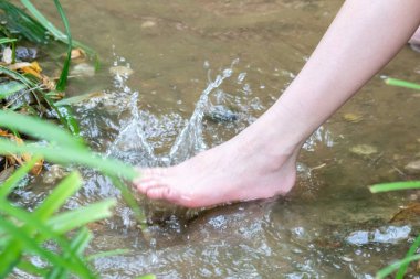 Küçük bir derede çıplak ayakla temiz suyla oynayan genç bir çocuk ayaklarını ve su kaynağını kullanarak ayak parmaklarını ve bacaklarını serinletiyor ve zen meditasyon atmosferindeki saf yaşam iksiriyle ferahlatıyor.