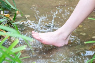 Küçük bir derede çıplak ayakla temiz suyla oynayan genç bir çocuk ayaklarını ve su kaynağını kullanarak ayak parmaklarını ve bacaklarını serinletiyor ve zen meditasyon atmosferindeki saf yaşam iksiriyle ferahlatıyor.