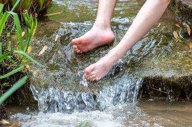 Küçük bir derede çıplak ayakla temiz suyla oynayan genç bir çocuk ayaklarını ve su kaynağını kullanarak ayak parmaklarını ve bacaklarını serinletiyor ve zen meditasyon atmosferindeki saf yaşam iksiriyle ferahlatıyor.