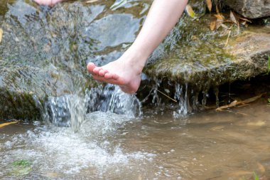 Küçük bir derede çıplak ayakla temiz suyla oynayan genç bir çocuk ayaklarını ve su kaynağını kullanarak ayak parmaklarını ve bacaklarını serinletiyor ve zen meditasyon atmosferindeki saf yaşam iksiriyle ferahlatıyor.