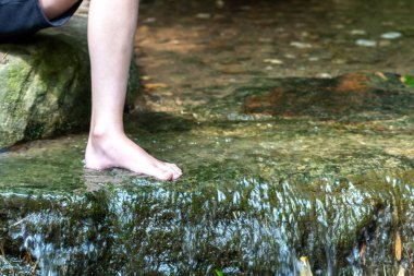 Küçük bir derede çıplak ayakla temiz suyla oynayan genç bir çocuk ayaklarını ve su kaynağını kullanarak ayak parmaklarını ve bacaklarını serinletiyor ve zen meditasyon atmosferindeki saf yaşam iksiriyle ferahlatıyor.