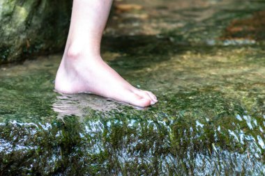 Küçük bir derede çıplak ayakla temiz suyla oynayan genç bir çocuk ayaklarını ve su kaynağını kullanarak ayak parmaklarını ve bacaklarını serinletiyor ve zen meditasyon atmosferindeki saf yaşam iksiriyle ferahlatıyor.