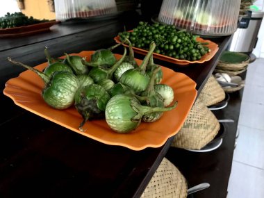 Turuncu bir tabakta sergilenen taze yeşil patlıcanlar, mutfak ortamında doğal sebzelerle birlikte, çiğ çiftlik ürünlerinin pişirilmeye ve günlük yemeklere hazır olduğunu gösteriyor..