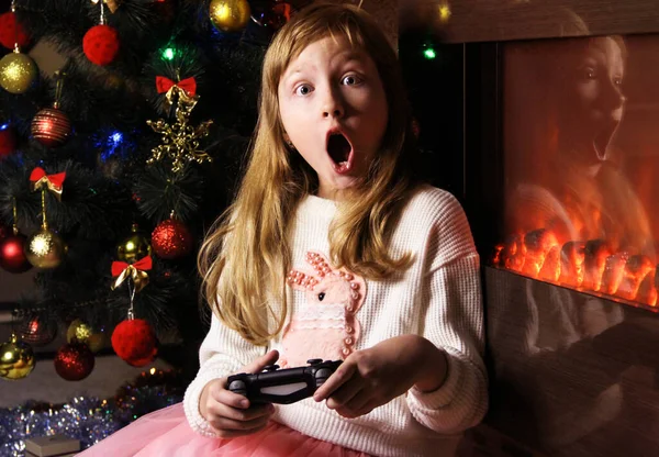 A girl with bright emotions plays a game console against the background of a Christmas tree and a fireplace. Player. New year\'s entertainment.