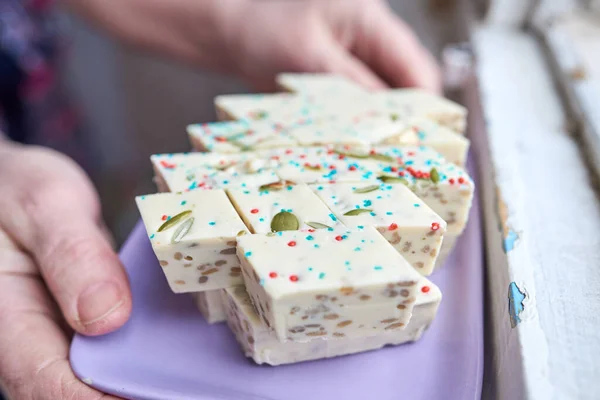 Samarcanda halva cortada en rombos con pistachos y anacardos y pasta azul y roja rematando en un plato rectangular púrpura en manos femeninas —  Fotos de Stock