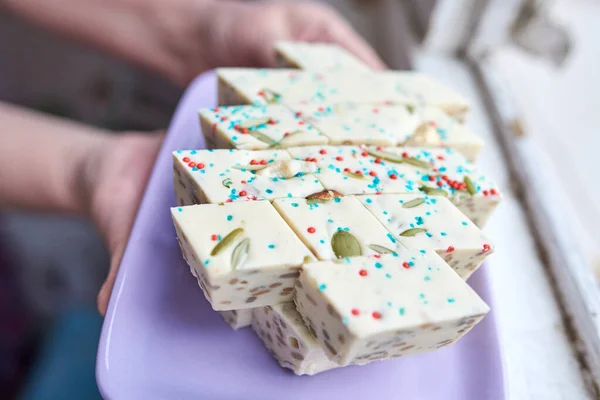 Samarcanda halva cortada en rombos con pistachos y anacardos y pasta azul y roja rematando en un plato rectangular púrpura en manos femeninas —  Fotos de Stock