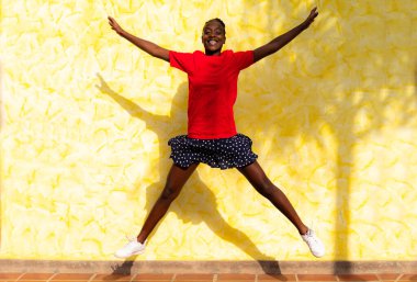 Gülümseyen genç bir kadın sarı arka planda havada zıplıyor. Dışarıda kutlayan ve dans eden mutlu Afrikalı kadın. Mutluluk, özgürlük, hareket ve insanlar kavramı.