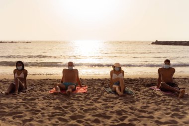 Korumacı yüz maskeli ve koronavirüs zamanında mesafeli çok ırklı arkadaşlar. Sıkıcı genç milenyum insanları yaz mevsiminde plajda oturuyorlar. Yeni normal yaşam tarzı ve sosyal uzaklık kavramı.