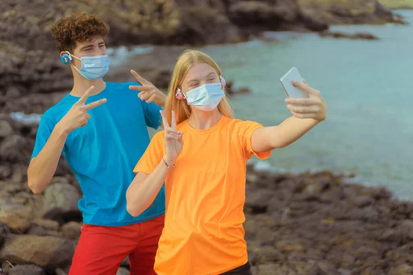 Young couple with protective mask listening music. Boy and girl taking selfie with smart phone,  portrait with rock beach scenery in background.