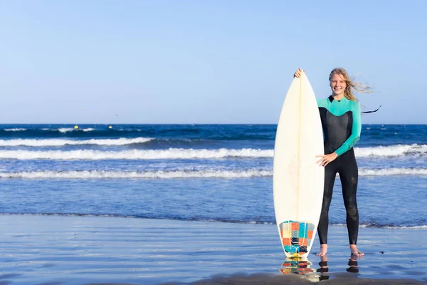 Blonde caucasian woman with a surfboard on beach. Smiling and posing with surf board near blue ocean in sunny day.