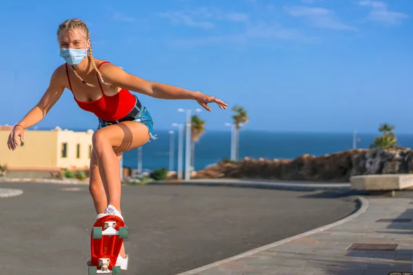 赤いTシャツと医療用マスクスケートでブロンドの髪を持つ美しい十代の女の子 市道でスケートを練習する女性スケーター スポーツとコロナウイルスの概念 — ストック写真