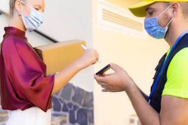 Akıllı telefona koruyucu maske takan beyaz kadın teslimatçıdan kutu, akıllı telefondan kadın imzası kabul ettikten sonra akıllı telefona imza attı. Teslimat ve koronavirüs konsepti.