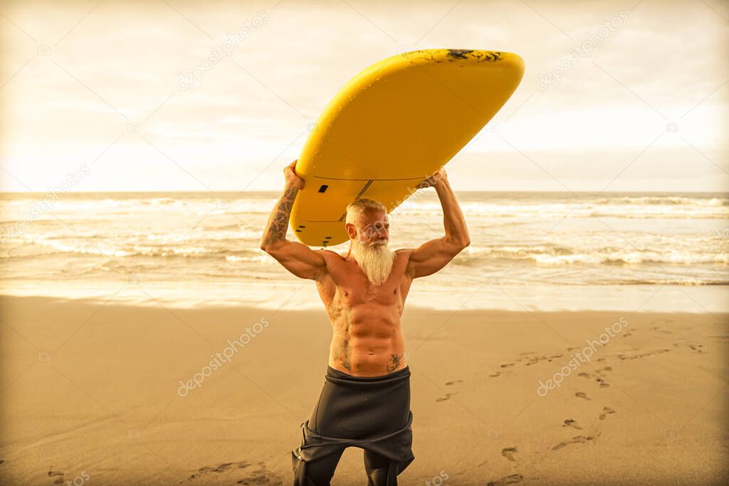 Happy fit senior having fun surfing at sunset time. Sporty bearded man training with surfboard on the beach. Elderly healthy people lifestyle and extreme sport concept.