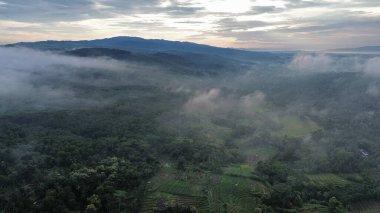 Sabahları arka plan olarak dağlar ve tepeler olan, çevresindeki tarım arazileri ve sabah sisi ile kırsal arazinin hava aracı görüntüsü.