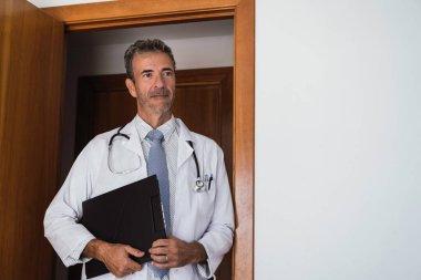 Kıdemli doktor mavi ceketli ve kravatlı. Kapıda duruyor. Kıdemli doktor elinde siyah bir klasörle kapıda duruyor. İlaç, sağlık hizmetleri