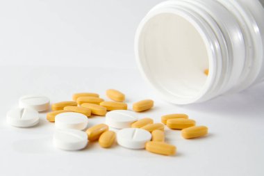 Beyaz ve sarı haplardan oluşan bir yakın çekim beyaz yüzeydeki ilaç şişesinden düşüyor.