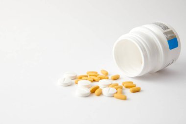 Beyaz ve sarı haplardan oluşan bir yakın çekim beyaz yüzeydeki ilaç şişesinden düşüyor.