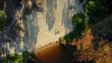 Nehir kenarında dinlenen insanların bulunduğu kumlu bir sahil manzarası. Drone güneşli bir günde kumsaldaki eski bir nehri rahatlatmak için havalanıyor. İnsanlar ve yazın nehir kenarında vahşi bir tatil.
