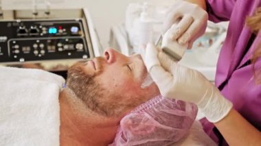 Güzellik uzmanı yüz işlemleri için elektriksel impulslar kullanıyor. Yüz bakımı için kozmetik işlemleri yapan bir spa salonundaki adam. Yetişkin adam spa salonunda mikroakım tedavisi görüyor..  