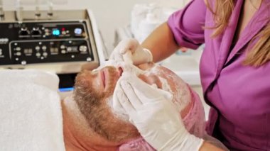 Kozmetik uzmanı erkek yüzünden maske çıkartıyor. Orta yaşlı bir adam yüz bakımı için kozmetik prosedürleri üzerine bir kaplıca salonunda çalışıyor..