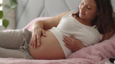 Hamile bir kadın çocuğuna bakarken kendini mutlu hissediyor. Genç yaşta hamile bir anne karnında bebeği tutuyor. Kadın hamilelik konsepti ve doğum öncesi bakım. Yavaş çekim 4K video.