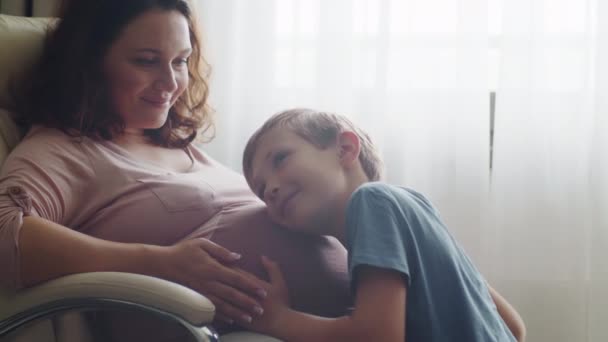 孩子们拥抱着并亲吻她母亲怀孕的肚子 快乐的儿子在家里的窗户附近摸着他怀孕的妈妈的大肚子 在期待新生儿的快乐家庭生活中 怀孕和分娩 — 图库视频影像