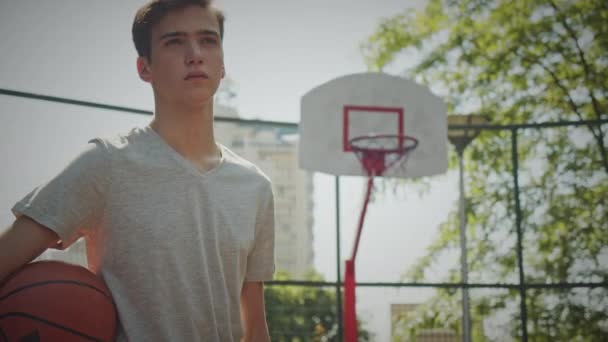 少年はバスケットボールコートに立ってバスケットボールをしている 10代の若者がボールを手に立ち バスケットボールのバスケットを背負って撮影した バスケットボール選手の自信に満ちた顔のビデオ 腕の下のバスケットボール — ストック動画