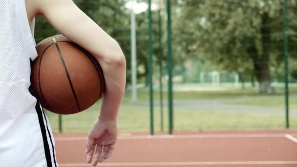 少年はバスケットボールコートに立って 腕の下でバスケットボールをしている 彼の手にボールとバスケットボールバスケット彼の前に立ってティーネージャーの背中のショットからビデオ — ストック動画