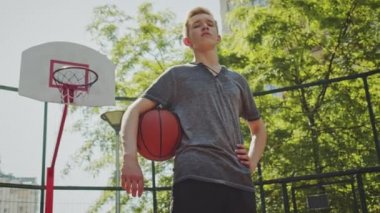 Basketbol sahasında duran ve elinde basketbol topu tutan bir çocuk. Elinde top, arkasında basketbol potası olan bir gencin sinematik çekimi. Kendine güvenen bir basketbolcunun videosu. Kol altında basketbol.