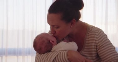 Genç bir anne oğlunu öpüyor ve onu elleriyle tutuyor. Sevgi dolu bir anne öpücüğünün yakın çekimi. Anne bebeğin yüzünü öpüyor. Anne bebeğine iyi bakıyor. Bebek bakışı. Mutlu aile. 