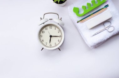 Beyaz çalar saat ve arka planda yeşil saksı bitkisi. Tırnak manikürü için beyaz, terli havlu, makas ve satıcı.