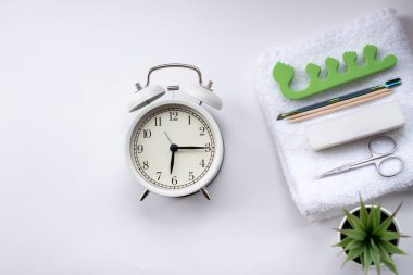 Beyaz çalar saat ve arka planda yeşil saksı bitkisi. Tırnak manikürü için beyaz, terli havlu, makas ve satıcı.