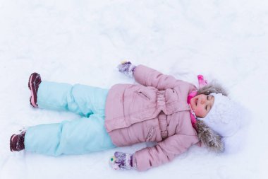 Mavi gözlü, beyaz şapkalı küçük bir kız karda yatıyor.