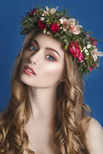 Vacker ung flicka med en blommig prydnad i håret på en blå bakgrund. Kvinna i kransen av blommor. Skönhet ansikte. Mode foto. Perfekt makeup. Hudvårdskoncept. — Stockfoto