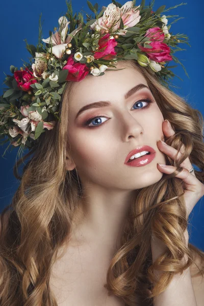Mooi jong meisje met een florale sieraad in haar haar op een blauwe achtergrond. Vrouw in krans van bloemen. Schoonheid gezicht. Mode foto. Perfecte make-up. Skin Care concept. — Stockfoto