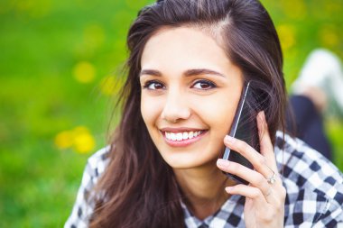 Portrait of a beautiful woman typing on the smart phone in a park with a green unfocused background.  Hipster woman texting in summer.