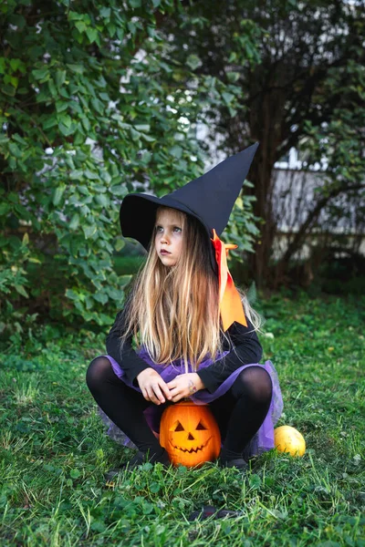 Zabawna dziewczynka w stroju czarownicy na Halloween siedzi na dyni Jack outdoor w parku — Zdjęcie stockowe