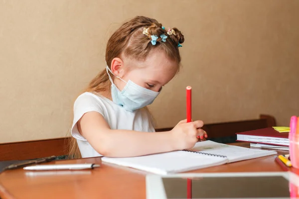 Cute little girl w masce ochronnej robi pracę domową w domu. Kształcenie domowe podczas kwarantanny. — Zdjęcie stockowe