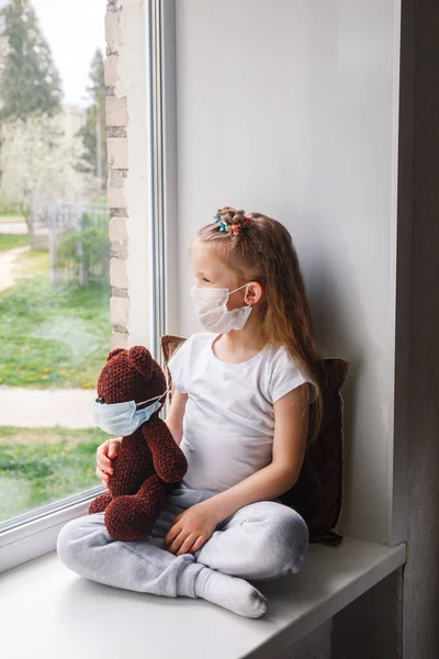 Chica solitaria en máscara de protección jugando con el juguete y sentado en la ventana. Concepto de cuarentena del coronavirus — Foto de Stock