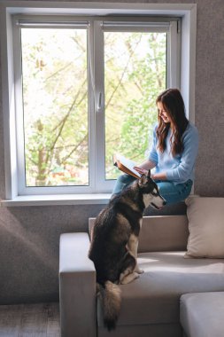 Evdeki pencerenin yanında kitap okuyan güzel genç bir kadın.